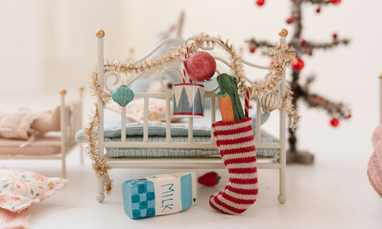 A small red-striped stocking filled with  fruits and veggies hanging from the end of the Maileg Micro Vintage Metal Bed. 